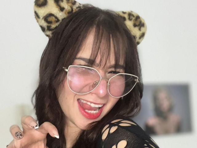 MelannyTaylorr, con cabello oscuro y ondulado, orejas de leopardo y gafas de color rosa, sacando la lengua, vistiendo una blusa de malla negra, mostrando su lápiz labial rojo y su anillo..