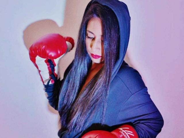 Roxanne Marquez, con cabello negro largo, guantes de boxeo rojos y una sudadera negra, sosteniendo los guantes, mirando hacia abajo, con lápiz labial rosa, iluminación púrpura suave, expresión tranquila, concentrada..