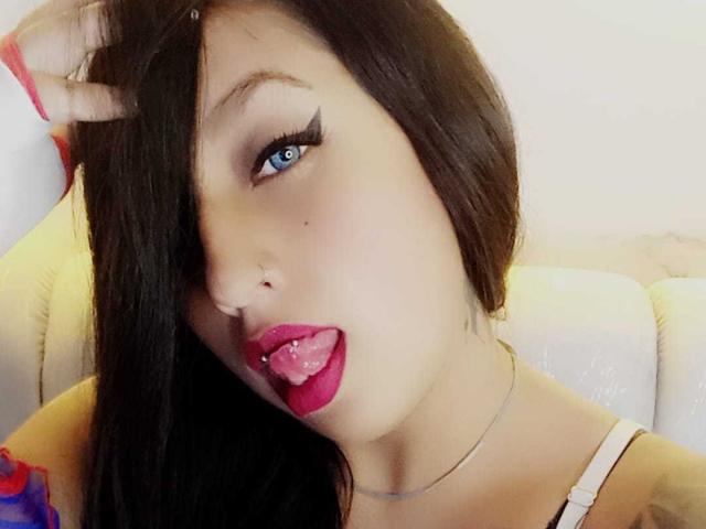 Roxanne Marquez, con cabello negro, ojos azules, delineador de ojos negro grueso, lápiz labial rosa, lengua afuera, collar de plata, mano en el cabello, correa blanca y un tatuaje discreto en el brazo izquierdo..