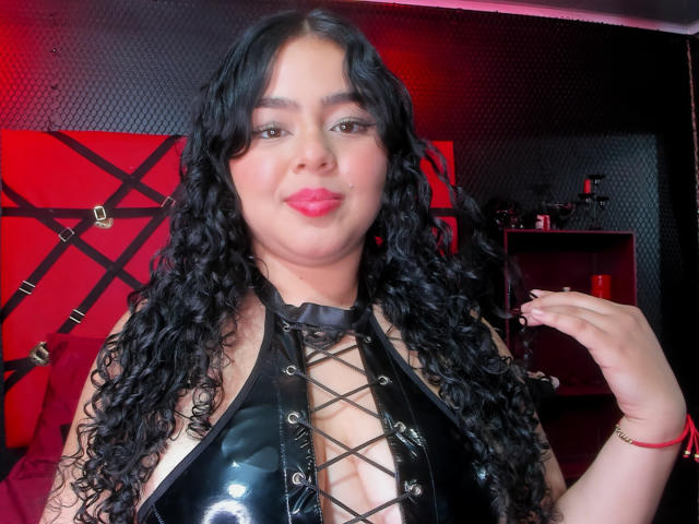 Apriel Keith a les cheveux noirs et bouclés, et elle porte une tenue en cuir noir brillante avec une fermeture à lacets sur le devant, qui dévoile sa poitrine. Elle sourit avec ses lèvres roses et touche ses cheveux..