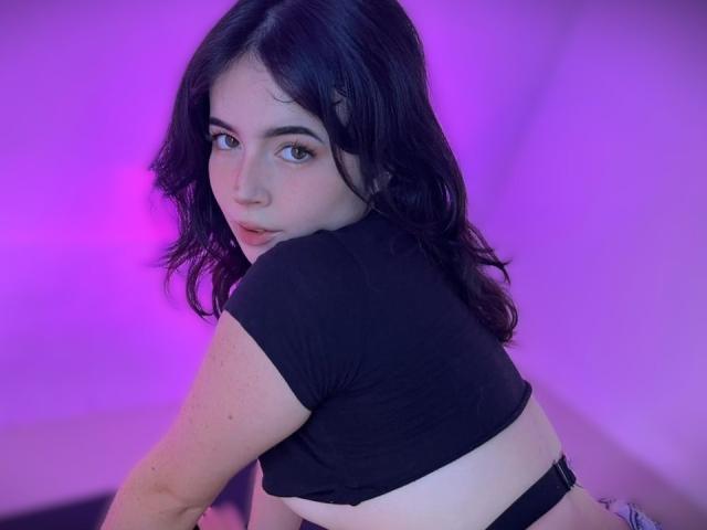 AliinaFontaine with wavy black hair, looking over her shoulder, wearing a black crop top, showing her back and black panties, in purple lighting, with a subtle smile.
