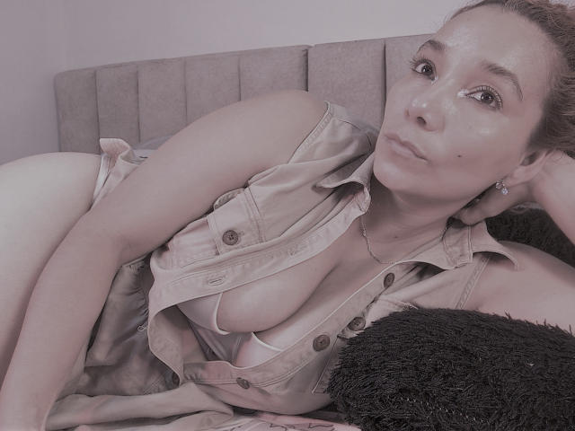 AmberDemons met bleke huid, kleine borsten en een lichte bruine tint, liggend op een bed, met een open beige shirt aan, waarbij een beetje van haar borsten zichtbaar is, kijkend naar de camera..