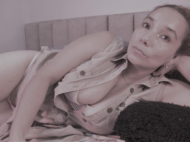 AmberDemons liggend op bed, met een beige overhemd dat open staat, waardoor haar grote borsten en een roze beha zichtbaar zijn, met een pluizig zwart kussen, kijkend naar de camera, met zachte roze verlichting..