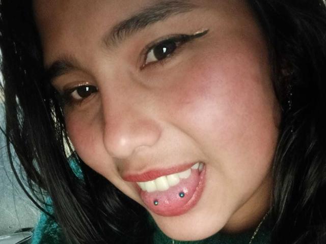GemmaBellini's face, dark hair, brown eyes, green shirt, smiling with tongue out, lower lip pierced with blue and black studs. She's got winged eyeliner and a cute, fresh-faced look.