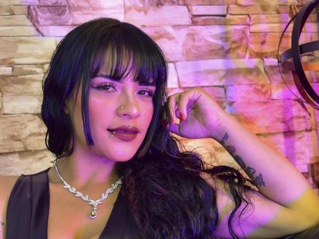 Mia Bellucci, con cabello negro, flequillo y reflejos morados, tiene un piercing en la nariz, un piercing en el labio y un tatuaje en el antebrazo derecho, y lleva una blusa negra y un collar con adornos..