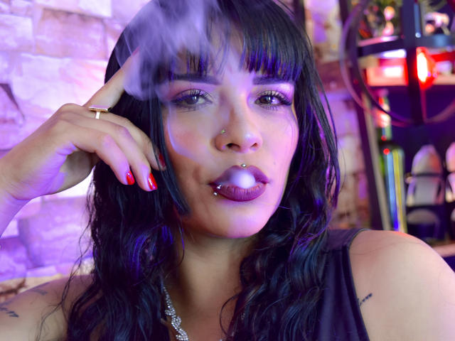Mia Bellucci, met zwart haar, paarse lippenstift en rode nagellak, die een joint rookt, heeft een neusring en een lip-piercing, draagt een zwarte top, in een paars verlichte omgeving..