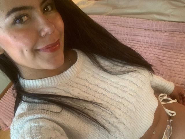 CatalinaReiyna with long dark hair, smiling, wearing a white knit sweater, showing her small tits, and a white string bikini bottom, sitting on a pink quilted bed.