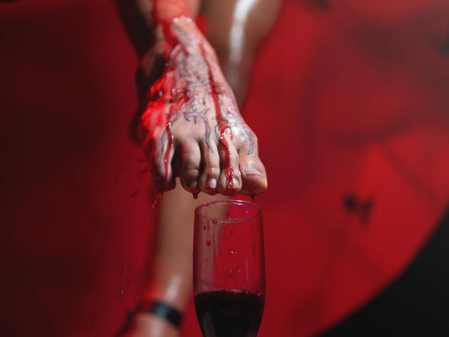 Kalyban's bloody hands grip her toes, which are above a glass of red wine. Blood splatters her tattoos and she wears a black bracelet. Red light.