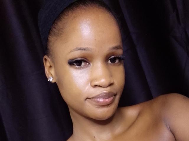 PieretteE has dark brown skin, a black headwrap, and is looking at the camera with a slight smile. She's wearing a nose ring and diamond earrings. Her tits are not visible.