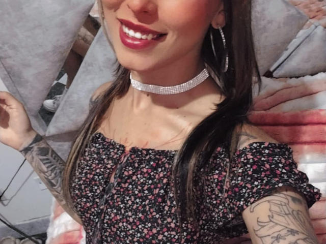 LunaLindaHot with red lipstick, floral dress, off-shoulder, sparkling choker, long brown hair, tattooed arm, smiling, hoop earrings, and visible cleavage.