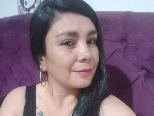 VivianMilf has black hair, pink lipstick, and hoop earrings, wearing a black tank top, showing a tattoo on her right shoulder, and smiling at the camera.