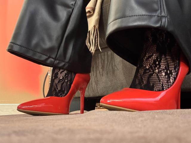 HannaBorbon's glossy red high heels with stiletto heels, black lace socks, and black leather pants, standing on a gray carpet. Beige scarf partially visible.