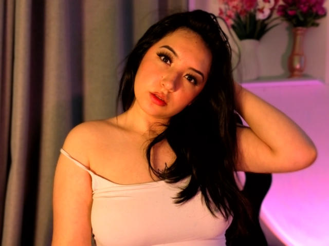 SusanMonroy with long black hair, pale skin, and red lips, wearing a white off-shoulder top, hand in hair, pink lighting, looking slightly pouty, pink flowers in background.