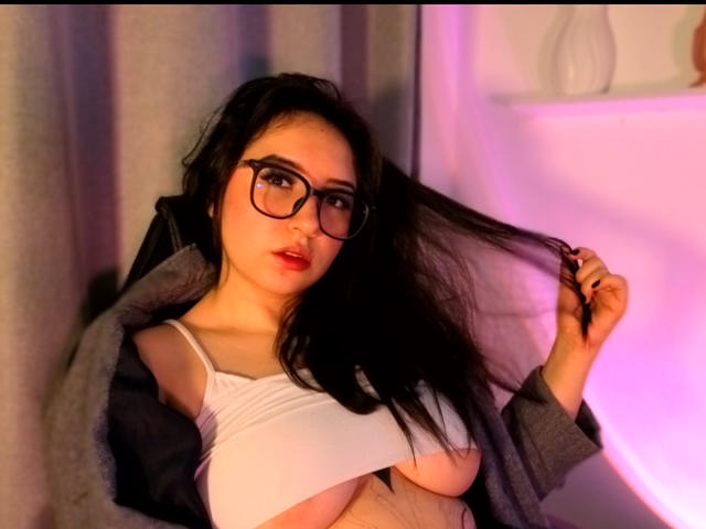 SusanMonroy with glasses, black hair, and red lipstick, holding hair, wearing open gray jacket, white top, exposing large tits, sitting in soft pink and purple light.