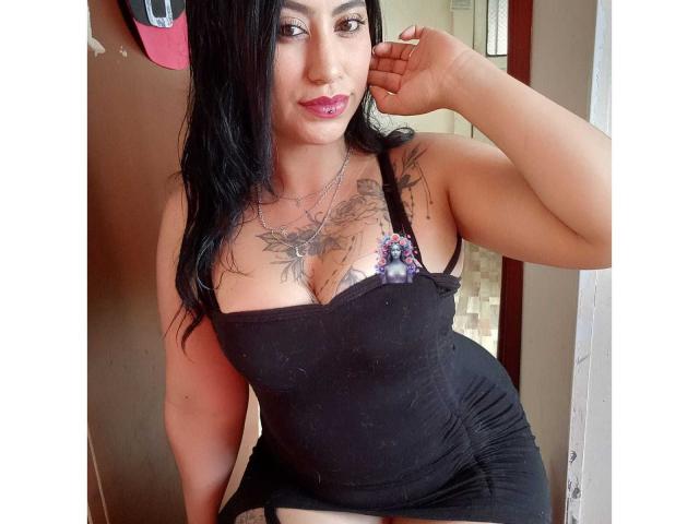 AlanaSweet69 with black hair, pink lipstick, black tank top showing her tits, tattoo on chest, black skirt, right hand touching cheek, standing in doorway, looking at camera.