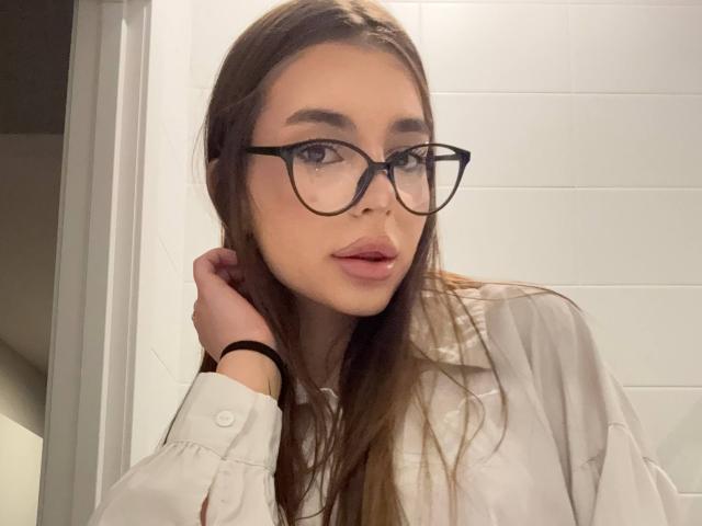 RoseleeVandiest with long brown hair, black-rimmed glasses, white shirt, and pink lips, holding her hair near a white tiled wall. Her ass is not visible.