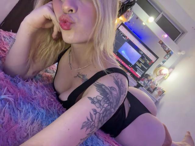 SammyViolet with blonde hair, pouty red lips, black lingerie, tattooed arm, and a fluffy pink-blue blanket, lying on her stomach, showing her ass.