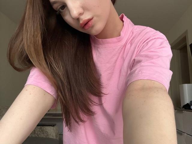 BaoRoebke in a pink tee, brown hair, pale skin, pouty lips, slightly tilted head, freckled arms, looking at camera, soft light, casual selfie.