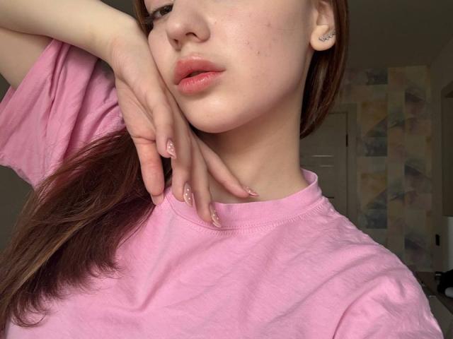 BaoRoebke with pink shirt, hand on cheek, pink nails, glossy lips, light skin, brown hair, subtle tattoos, soft lighting, slight smile, and a cute, relaxed facial expression.