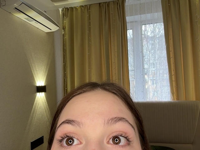 BaoRoebke's face, brown eyes looking up, with brown hair, visible forehead, and slight brow. Air conditioning unit above, beige curtains, and window with outside trees in background.