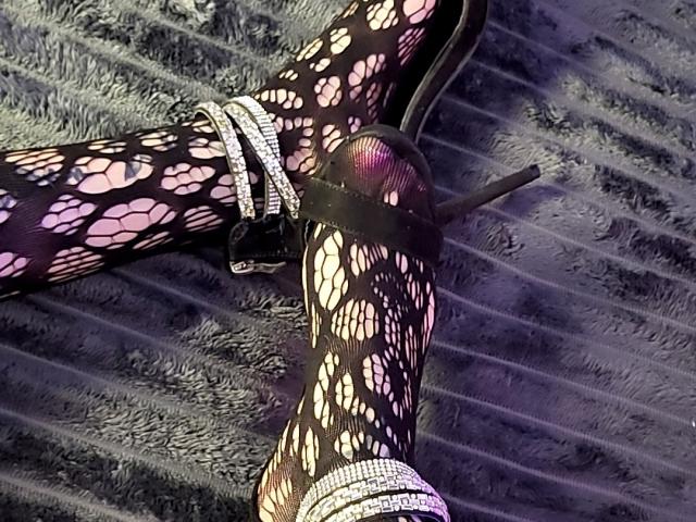 ElixirShanna's legs in black, pink, and white patterned fishnet stockings, with shiny silver straps and black high heels, lying on a textured, grey surface.