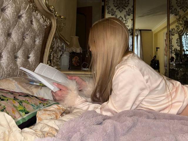 DannieCudney with long blonde hair, in pink satin pajamas, lying on a bed, reading a book, wearing fluffy wrist cuffs, with a floral pillow and purple blanket.