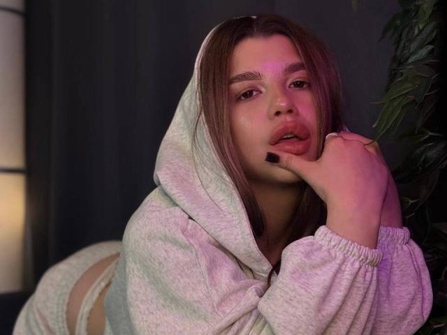 DannieCudney with long brown hair, gray hoodie, and matching pants, biting her finger with her hand on her cheek, pinkish-purple light on her face, looking seductive.