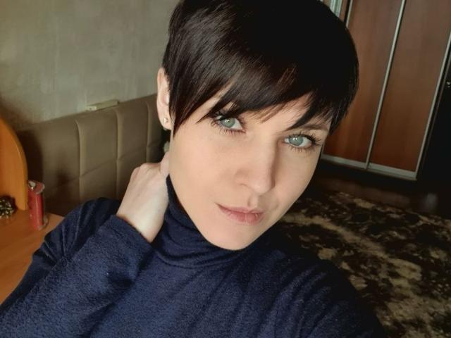 WenTina with short black hair, blue eyes, and a slight pout, wearing a dark blue turtleneck, has her hand on her neck, looking directly at the camera.