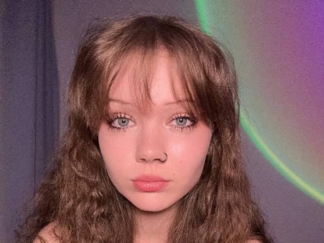 FannyEnsell has wavy brown hair, blue eyes, and pink lips with a nose ring. She looks directly at the camera with a neutral expression. Her tits are not visible.