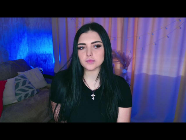 AdeleFelloy with long black hair, wearing a black shirt and cross necklace, has full eyebrows, pink lipstick, and a neutral expression, standing in a room with blue lighting.