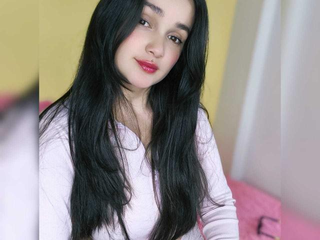 NataliaRivas with long black hair, pink lipstick, and a light purple shirt, smiling softly. Her tits are partially visible under the shirt. Her ass is not shown.