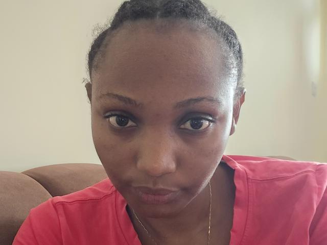 SerenityMoore with dark skin, braided hair, and brown eyes, wearing a red shirt, has a serious expression and a gold necklace, her pouty lips are slightly puckered.