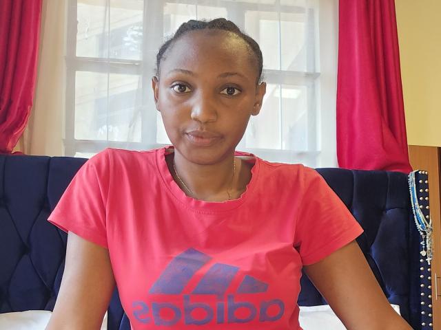 SerenityMoore with dark skin, short braided hair, wearing a pink Adidas t-shirt, sitting on a blue upholstered bed, red curtains in the background.