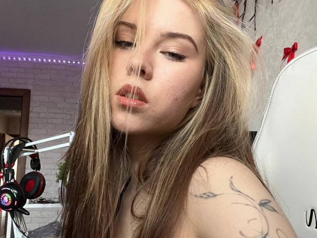 YolandoMysak with long blonde and brown hair, subtle makeup, and a tattoo on her right shoulder, looking down with slightly parted pink lips. Headphones on a stand in the background.