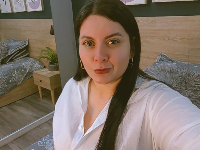 RachelCarlton with long black hair, white shirt, and red lipstick, has medium tits, and a cute, confident smile, standing in a bedroom with wooden furniture and a mirror.