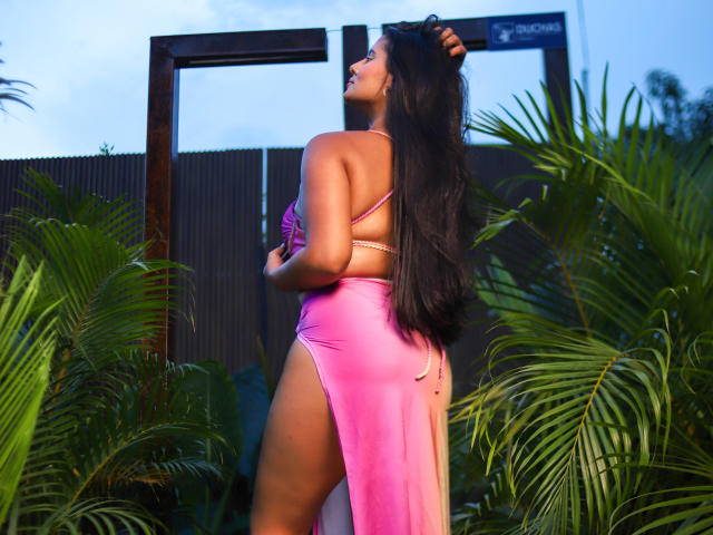 AnitaSage in a pink dress with high slit, showing her ass, long black hair, standing amidst green palm leaves, looking up, hand in hair, sunset light on her skin.