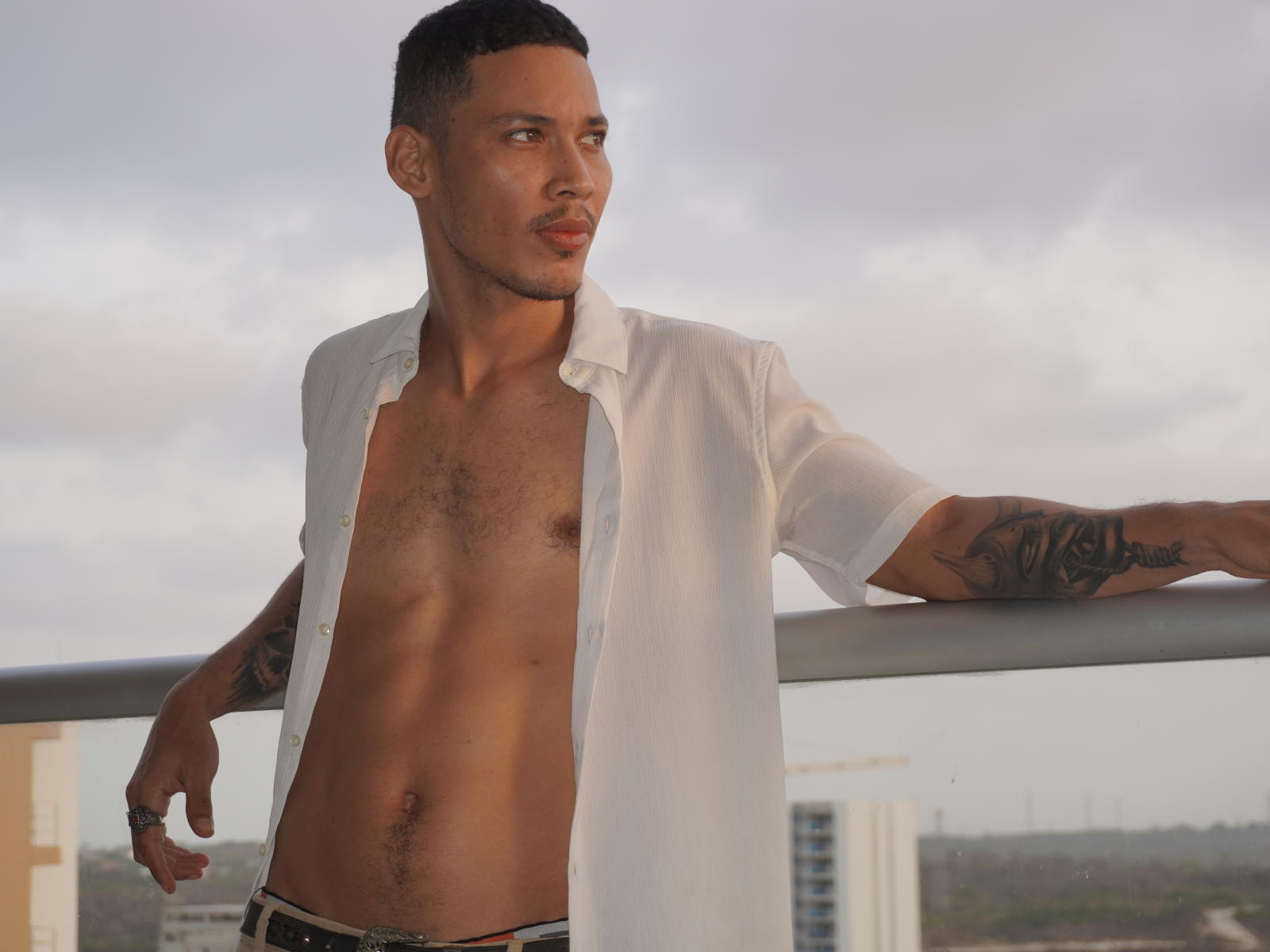RuiMaddoox, met een gespierd, behaard borstbeeld, een open wit shirt, tatoeages op zijn armen en zwart ondergoed, leunt tegen een balkon en kijkt naar de zijkant..