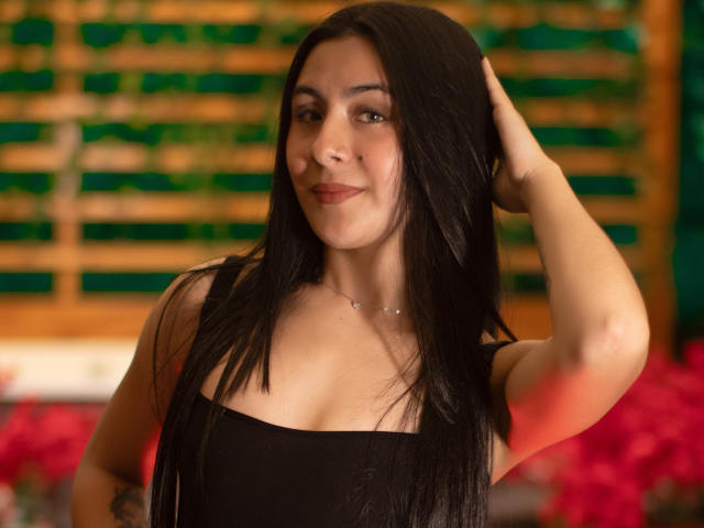 ChloeXk mit langem, schwarzem Haar, die leicht lächelt, trägt ein schwarzes Tanktop, hat eine Hand im Haar, ist mittelgroß, hat helle Haut, braune Augen, dezentes Make-up und eine Halskette, vor einem roten Hintergrund..