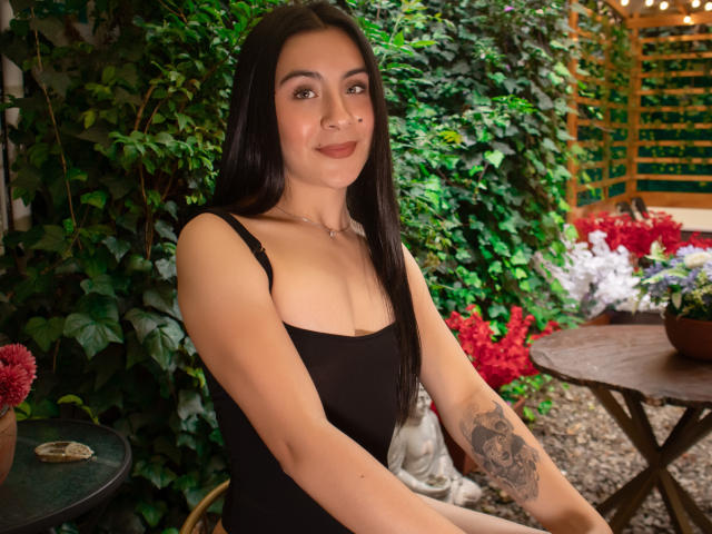 ChloeXk a de longs cheveux noirs, une peau mate, de petites seins, porte une robe noire, a un tatouage sur le bras droit, elle sourit et est assise à l'extérieur, près d'une table en bois avec une petite statue..