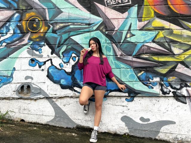MaraCortes with long black hair, wearing a purple shirt, black shorts, and white sneakers, standing against a colorful graffiti wall, lifting one leg and holding a cigarette.