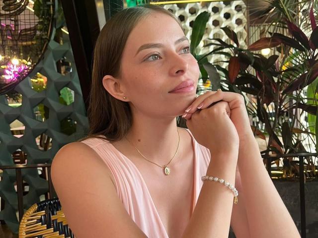 AnnaFer with brown hair, pink dress, gold necklace, and bracelet, thinking with her chin on her hand, showing her tits slightly, green eyes, and pink lips, in a plant-filled cafe.