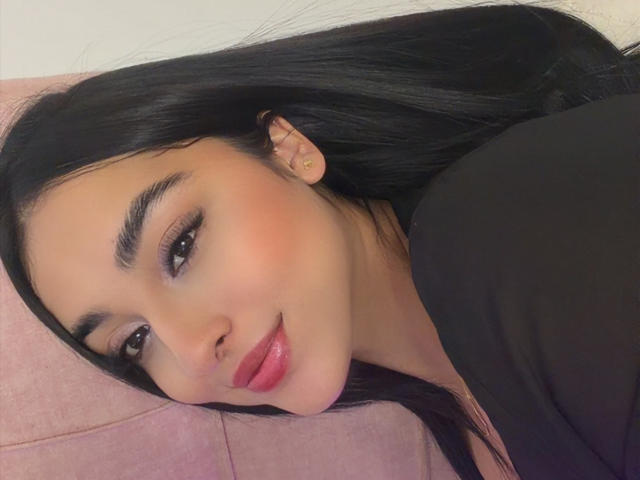 EmmaWaltter's face, black hair, pink lipstick, and dark eyes are visible, lying on a pink surface with a black top, small ear earring, and a slight smile.