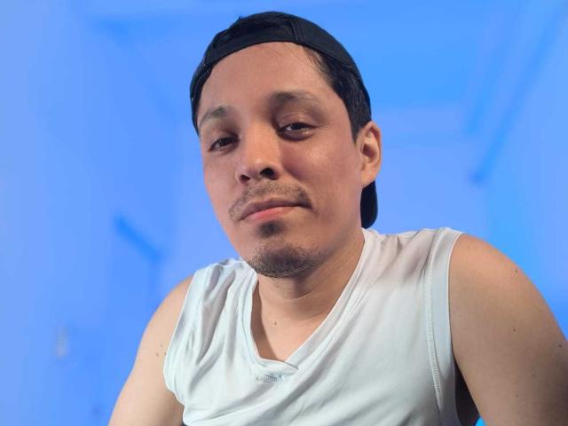 LukaForge, a chubby Asian dude with a small goatee, wearing a black backwards cap and a white tank top, looking slightly bored. Blue background.