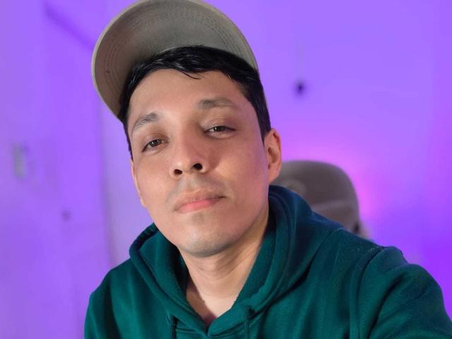 LukaForge with a neutral expression, wearing a green hoodie and gray cap, has a slight smirk and short black hair, against a purple-lit background.