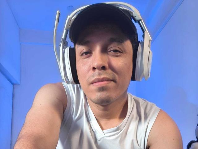 LukaForge with a medium build, wearing a white sleeveless shirt, and large white headphones, has a slight stubble, and a neutral expression, looking down at the camera.