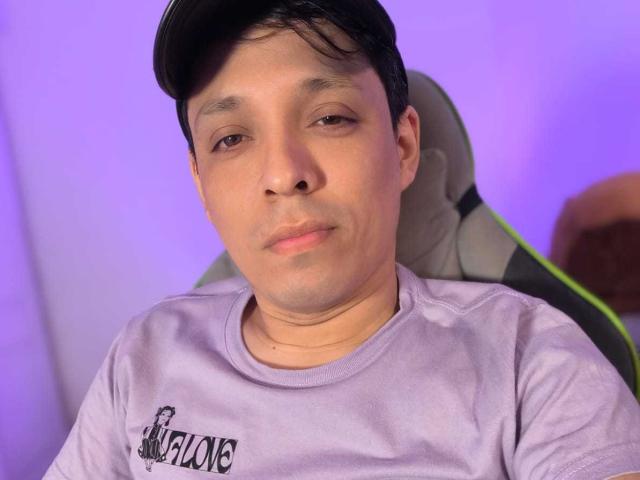 LukaForge with short black hair, light skin, slight smile, wearing a purple shirt, sitting in a gray chair against a purple-lit background. Boobless, ass unseen, no visible genitals.