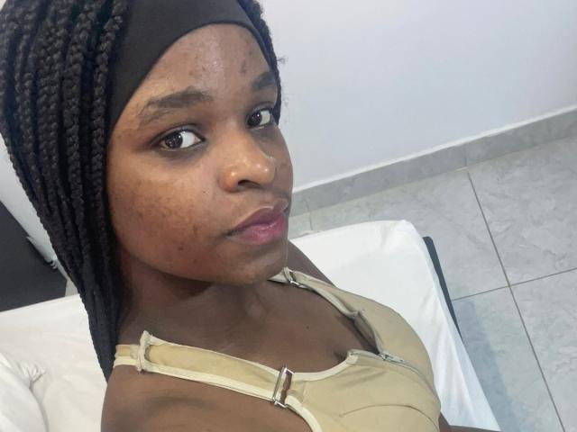 RenataWalker, dark-skinned with braided hair, wearing a black headscarf and beige tank top, showing freckles on her face, with a neutral expression and pink lips.
