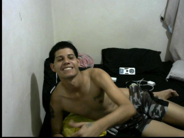 JackFox69, shirtless with black hair, smiling while lying on a bed, wearing camouflage shorts, holding a yellow pillow, with a black pillow and white wall in the background.