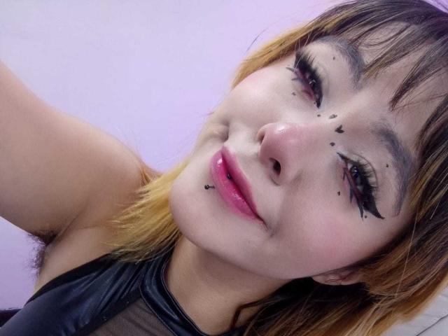 Le visage d'IvonnySakura, avec des points de khôl noir sur sa peau, du rouge à lèvres rose et des cheveux dégradés marron-jaune, portant un haut en maille noire, souriant à l'appareil photo..