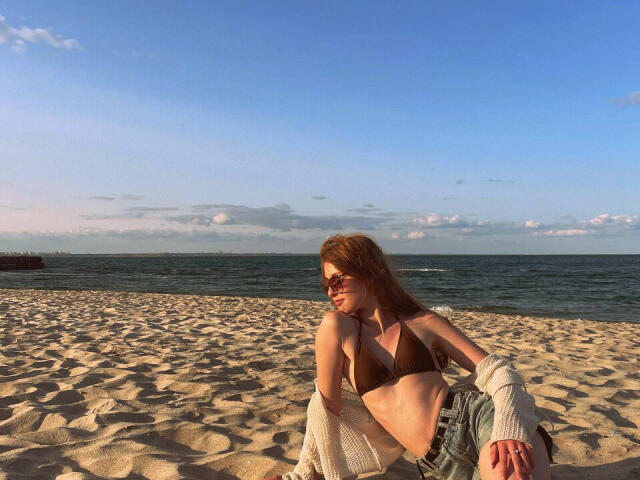 Nikollia con seni piccoli marroni, in un bikini marrone, seduta su una spiaggia sabbiosa, che indossa occhiali da sole, pantaloncini di jeans e un maglione bianco, con l'oceano e il cielo blu sullo sfondo..